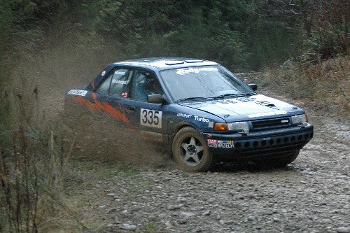 a dark blue mazda protege kicks up rocks on a gravel rally stage